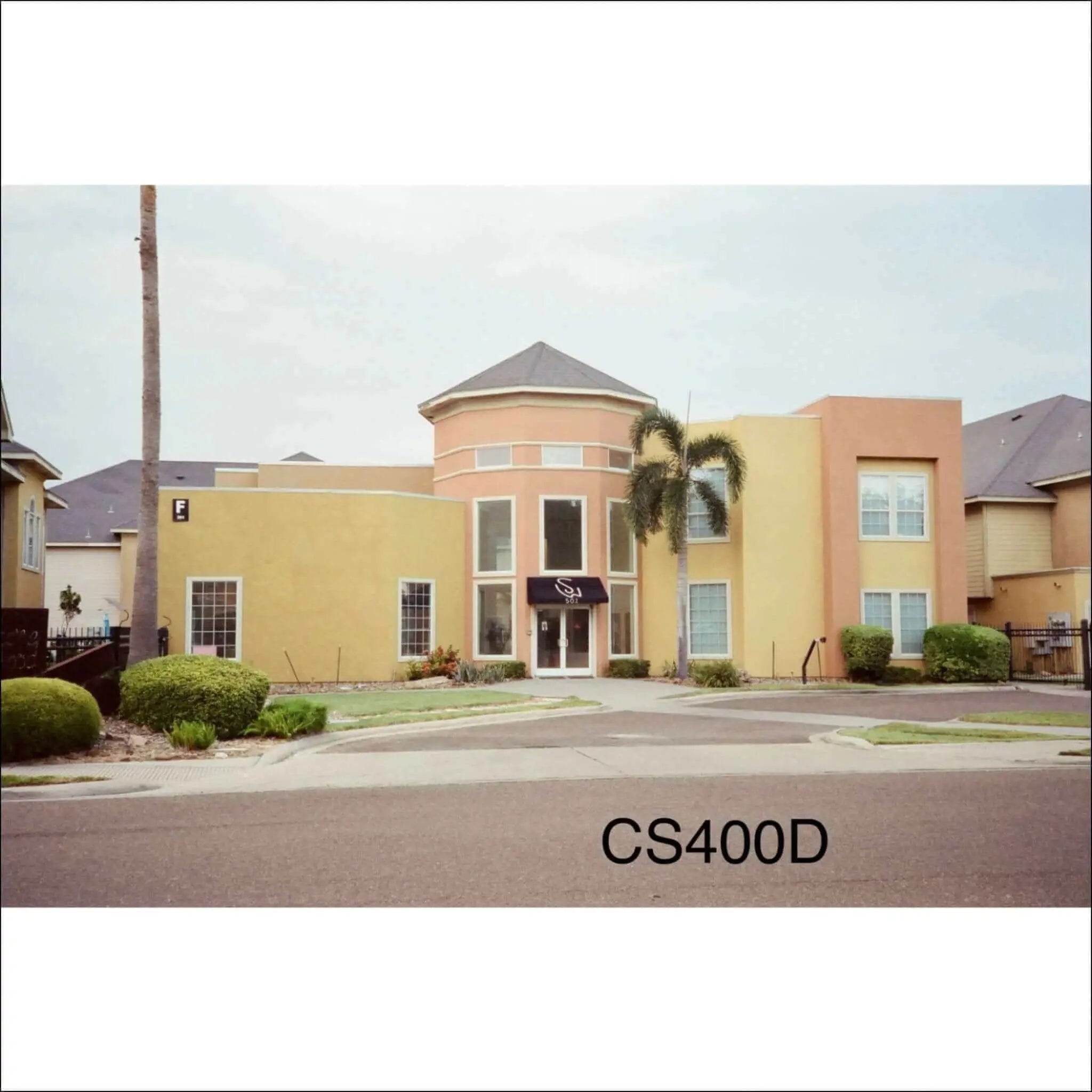 The image shows a two-story building with peach and yellow stucco walls, large windows, and a dark awning over the entrance.