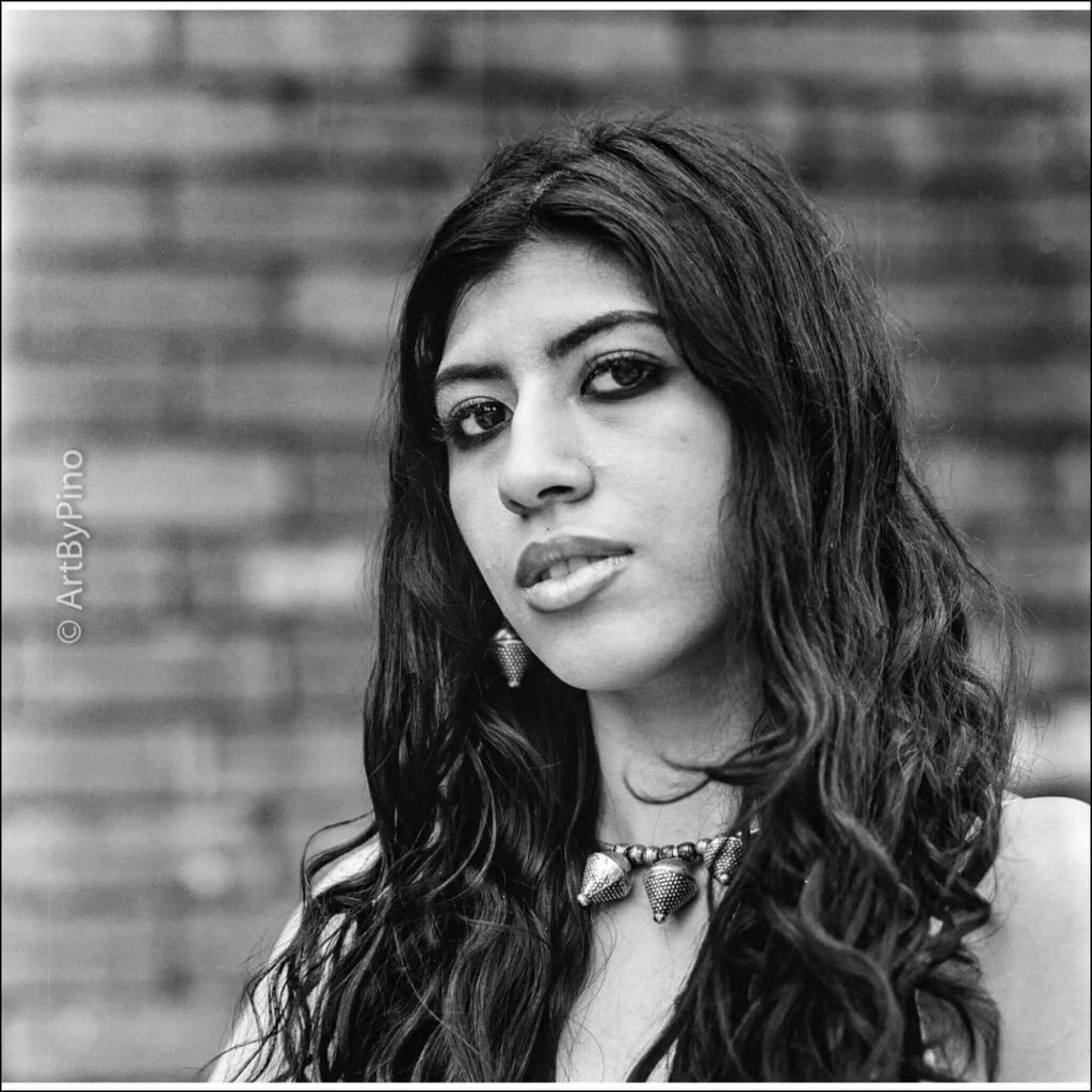 A black-and-white portrait of a woman with long, wavy hair wearing an ornate metallic necklace.