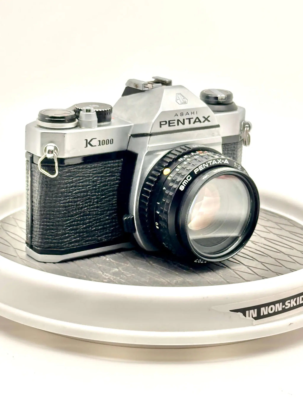 Silver asahi pentax k1000 film camera with black textured leather grip and a matching 50mm f/2.8 lens, resting on a non-slip surface.